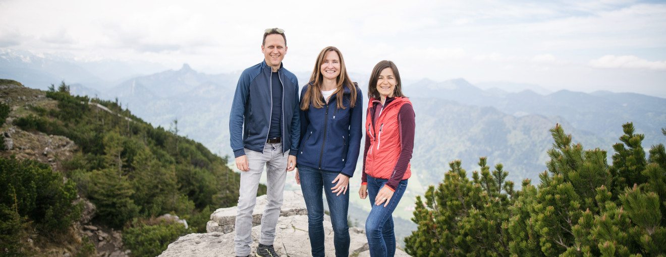 Ein Mann und zwei Frauen stehen lächelnd auf einem Berggipfel